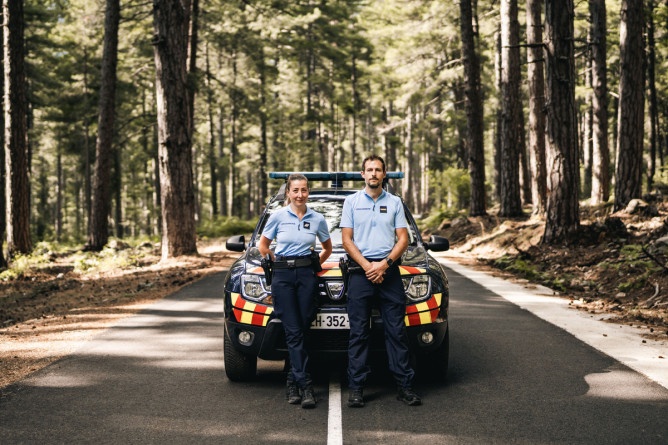 Agrandir l'image Deux jeunes gendarmes se tiennent debout devant leur véhicule de fonction, stationné sur une route au milieu d'une forêt.