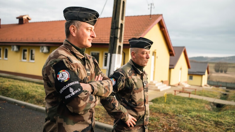 Agrandir l'image Deux prévôts français, de trois quart droite, en treillis arborant le brassard de la prévôté au bras droit. Le plus à gauche a les bras croisé et l'autre les mains sur les hanches. En arrière-plan les bâtiments jaune recouverts de tuiles du camp roumain