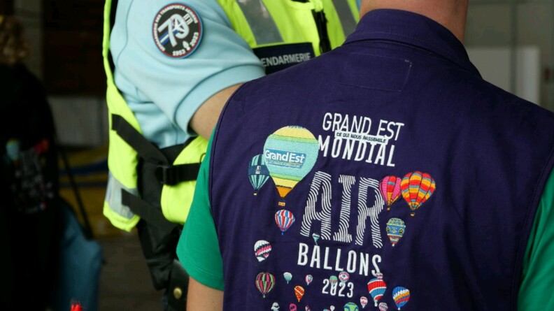 Agrandir l'image Au premier plan, un personnel de l'événement avec un polo avec pour inscription "Grand Est Mondial Air Ballons 2023 France". Autour de cette inscription des ballons. Au second plan, un gendarme de la GTA