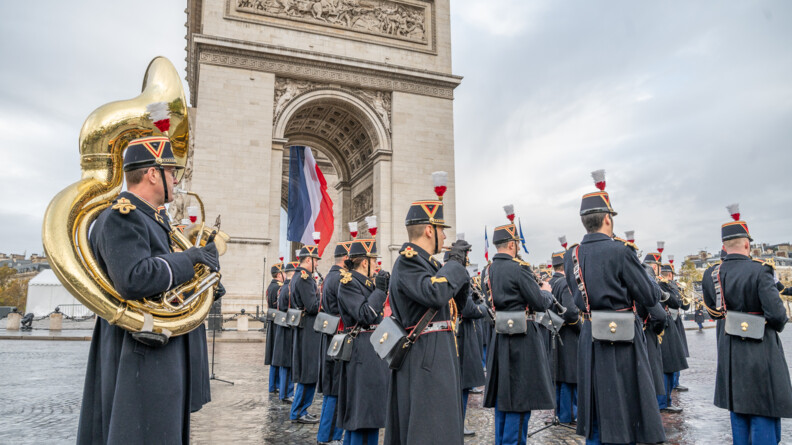 Des musiciens de la Garde républicaine en tenue, vêtus d'un long manteau noir, pantalon bleu marine, et du traditionnel chapeau à plumes, jouent divers instruments de musique sur le sol pavé de la place Charles de Gaulle à Paris. Derrière eux, se trouve l'Aec de Triomphe. Le ciel est gris.