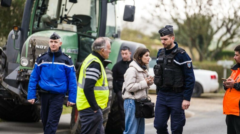 Deux gendarmes départementaux en tenue de service courant marchent au milieu des agricutleurs venus manifester. En fond, l'on voit un tracteur et une voiture.
