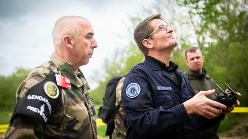 Deux gendarmes en buste, à gauche un prévôt en treillis, de profil, et à droite un scientifique de l'Institut de recherche criminelle de la gendarmerie en combinaison bleue, avec une manette de drône en mains, regardant vers le drône