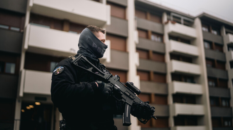 Un gendarme se tient debout, de biais par rapport &agrave; l'objectif, devant un immeuble d'habitations. Il tient un fusil contre son corps. Son visage est partiellement recouvert d'un cache-nez.