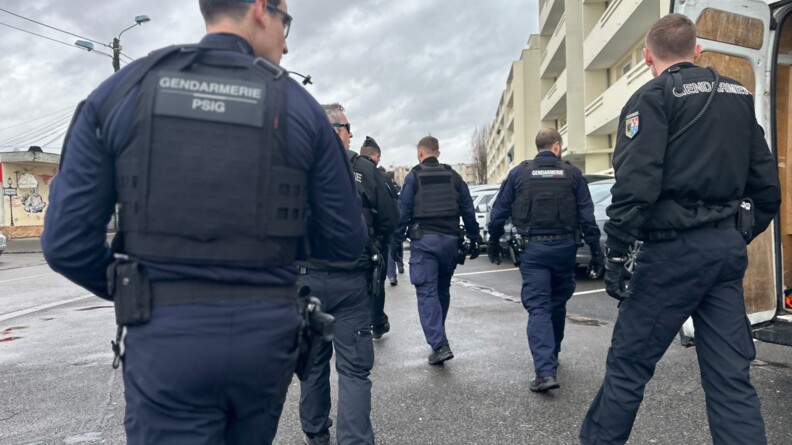 On aper&ccedil;oit de dos plusieurs gendarmes en tenue avan&ccedil;ant dans la rue, le long d'un immeuble d'habitation en b&eacute;ton blanc. Le ciel est gris.
