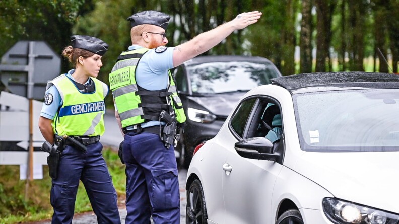 A gauche, deux gendarmes, une femme à gauche les mains dans le dos et un homme à droite indiquant une direction avec son bras droit, à un véhicule blanc vu de face. Derrière celui-ci, une voiture noire. En arrière-plan la forêt