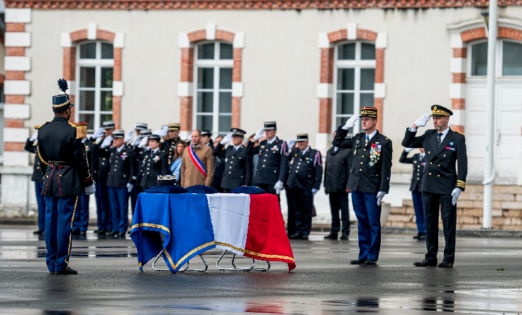 Agrandir l'image Des militaires saluent un cercueil recouvert du drapeau français. Au milieu d'eux, se trouve un élu, vêtu d'un manteau beige et de l'écharpe tricolore
