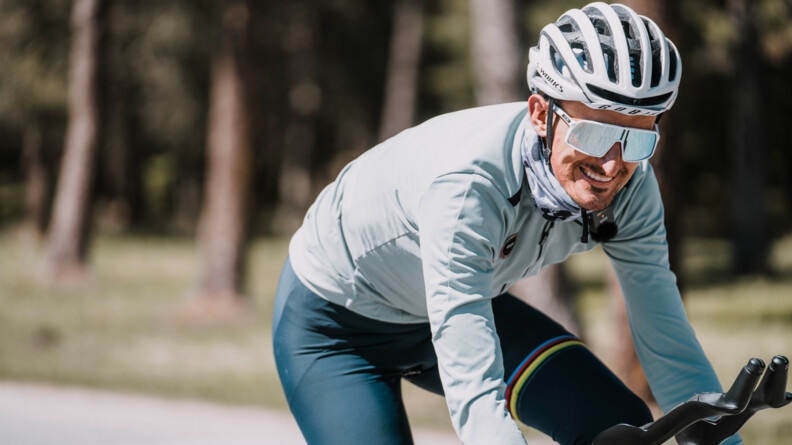 Un cycliste, avance, souriant, sur son v&eacute;lo, en regardant l'objectif. V&ecirc;tu d'une tenue bleue, il porte un casque, des lunettes de soleil blanches et des gants noirs.