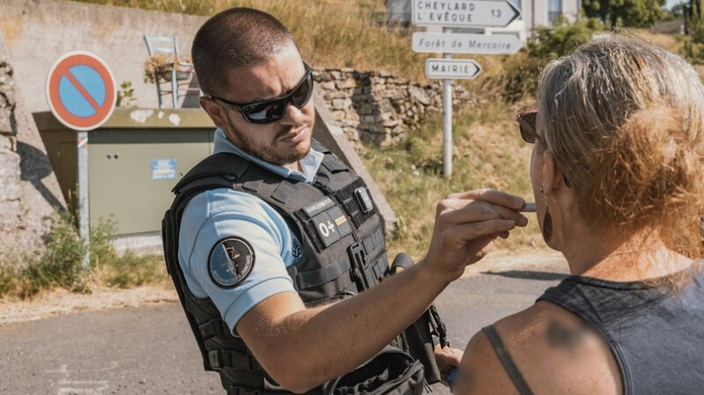 Un gendarme r&eacute;alise un test salivaire sur une femme, laquelle se tient debout, de dos. La sc&egrave;ne se d&eacute;roule en journ&eacute;e, sur une route de campagne.