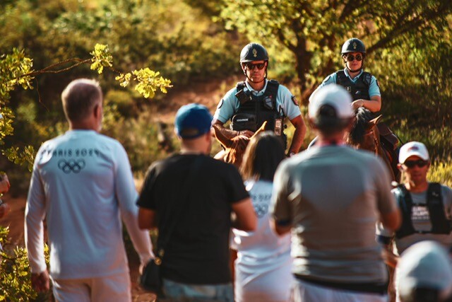 Au premier plan, quatre personnes de dos, dont deux portant un tee-shirt blanc siglé Paris 2024. Au second plan, à droite, un homme vu de face portant une casquette blanche et des lunettes de soleil, suivi par deux cavaliers de la gendarmerie sur leur monture, dont on n'aperçoit que les oreilles et le sommet de la tête.