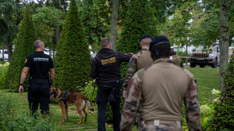 Qautre hommes de dos marchant dans une allée extérieure au milieu d'arbres. Parmi eux, un maître chien tient en laisse son malinois.