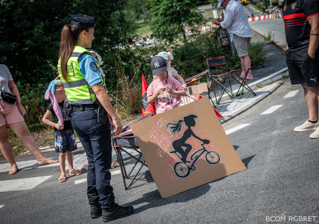 Agrandir l'image Une gendarme en uniforme, avec un gilet tactique jaune fluo et une queue de cheval, vue de trois-quarts dos, parle avec une enfant sur le bord de la route avec une pancarte d'encouragement pour une cycliste. D'autres spectateurs se trouvent à proximité au bord de la route.