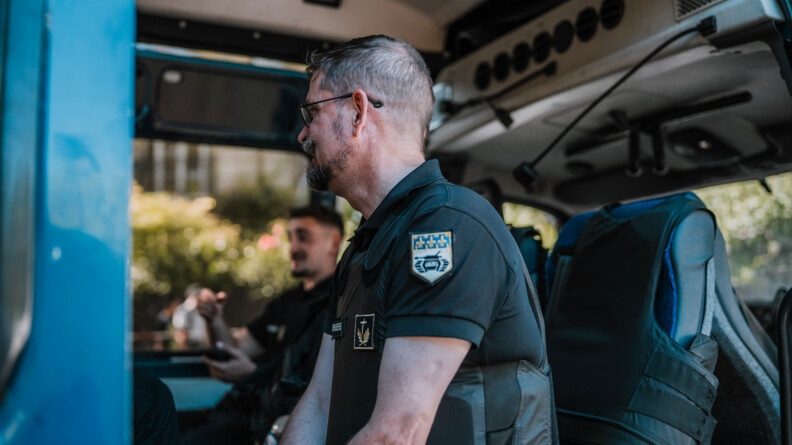 Un aumônier de gendarmerie de profil gauche assis à la porte d'un véhicule. EN flou au fond, ungendarme assis dans le sièeg