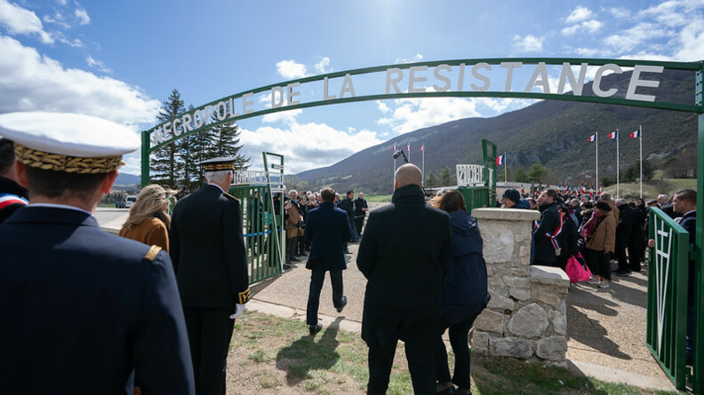 Agrandir l'image Des hommes et des femmes pénètrent dans un lieu, passant sous un porche vert sur lequel est inscrit "Nécropole de la Résistance". Le ciel est bleu et parsemé de nuages blanc.