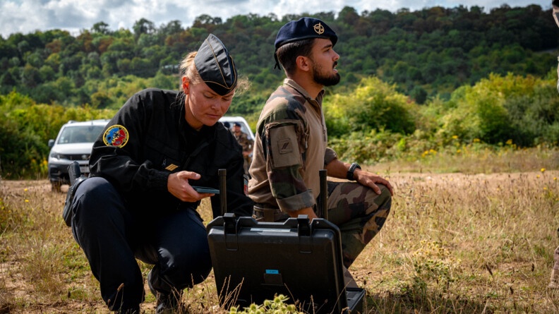 Une femme gendarme, vêtue d'un uniforme noir, se tient accroupie aux côtés d'un militaire. Une malette noire ouverte, est posée au sol, sur la pelouse, à leurs pieds. La femme tient un smartphone dans sa main droite, dont elle regarde l'écran. Deux véhicules, ainsi que des militaires, apparaissent en second plan.