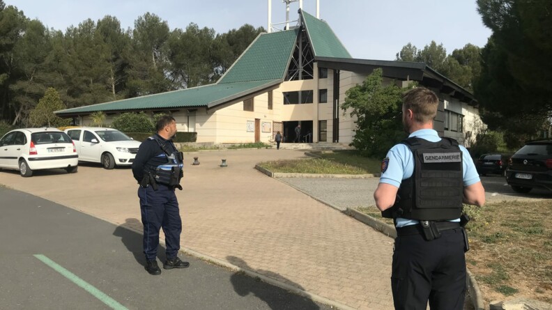 Au premier plan à droite un gendarme de dos; et un policier municipal de profil un peu plus loin. Au second plan à droite et à gauche des véhicules garés. Au milieu une allée pavée menant vers une église moderne au toit vert. En fond et de part et d'autre de l'église, des épineux.