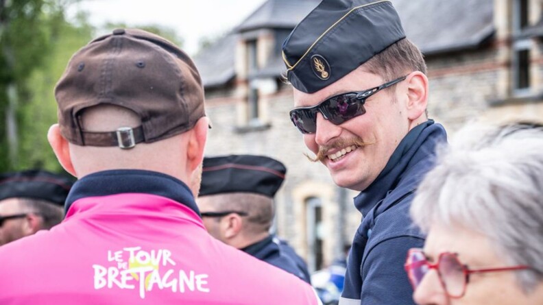 Un homme de dos, vêtu d'un polo rose portant le logo 'Le Tour de Bretagne" discutant avec un gendarme arborant un grand sourire.