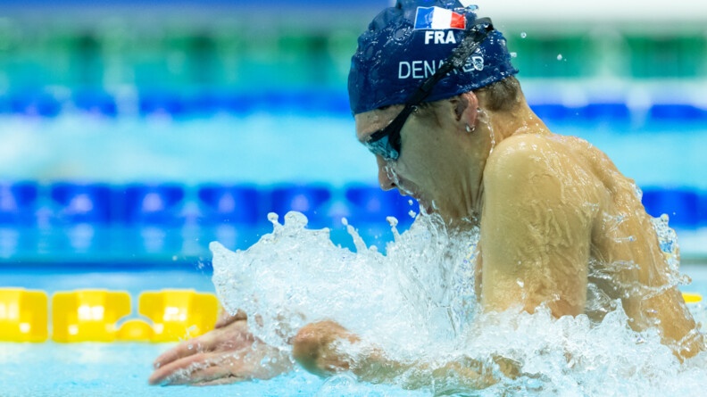Agrandir l'image De profil, sortant de l'eau faisant une vague autour de lui, une garçon portant des lunettes de natation noires et un bonnet bleu marine siglé d'u drapeau français et du nom Denayer, s'apprête à replonger dans l'eau
