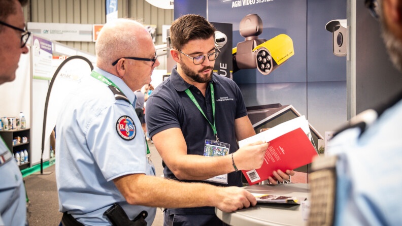 Un gendarme référent sûreté de profil droit échnage avec un homme en polo bleu marine qui lui montre une brochure. Devant eux en fond un kakemono pour de la video surveillance