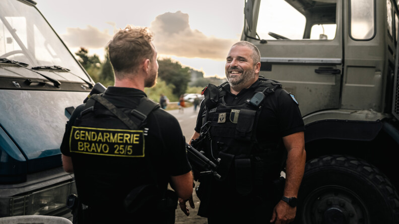 De dos un gendarme mobile dont la chasuble revet l'inscription en jaune "Gendarmerie Bravo 25:5", porteur d'une arme d'épaule. Face à lui, légèrement sur la droite, un aumônier s'adresse à lui, souriant. A gauche du gendarme se trouve l'avant d'un véhicule bleu et derrière l'aumônier celui d'un camion kaki