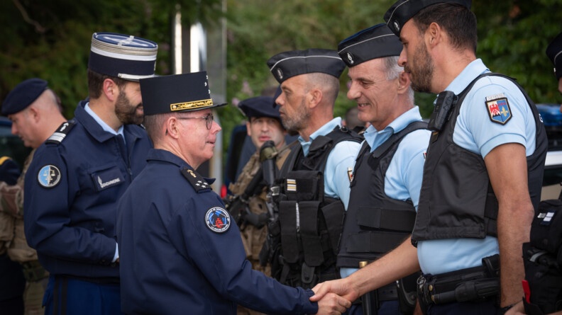 Le Major G&eacute;n&eacute;ral de la Gendarmerie Nationale serre la main &agrave; un gendarme. D'autres militaires de la gendarmerie et de l'arm&eacute;e de Terre, se tiennent debout &agrave; leurs c&ocirc;t&eacute;s