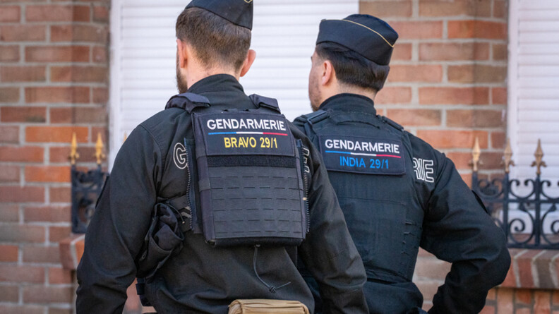 La photo repr&eacute;sente deux gendarmes avan&ccedil;ant dans la rue au c&ocirc;t&eacute;s des carnavaleux, de dos, devant une maison en briques rouges.