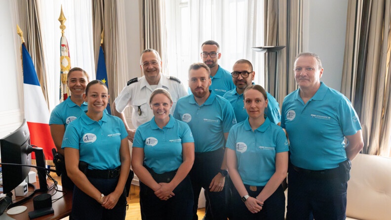 Agrandir l'image Dans un bureau, le directeur général de la gendarmerie pose au milieu de huit gendarmes vêtus d'un polo bleu turquoise. A leur gauche, on aperçoit un bureau et un grand drapeau français