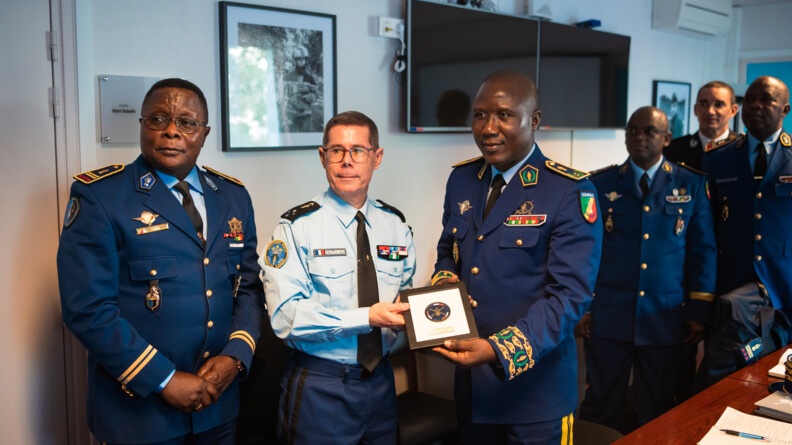 Agrandir l'image Dans une pièce fermée, un homme de la gendarmerie nationale se tient debout entouré de quatre gendarmes congolais. Le gendarme français tient un acdre carré contenant un insigne, avec l'un des gendarmes congolais. Derrière eux, se trouve un grand écran télévision ainsi qu'un cadre contenant une photographie. Au premier plan, on aperçoit une table sur laquelle sont posés des cahiers et un stylo.