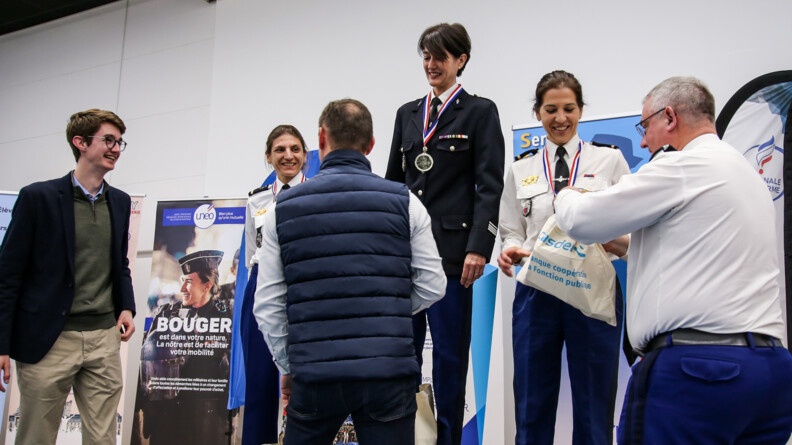 Agrandir l'image Remise des prix à trois gendarmes féminines sur le podium; trois hommes (un à gauche de profil, un de dos et un à droite de profil en tenue gendarmerie) leur remettent des lots