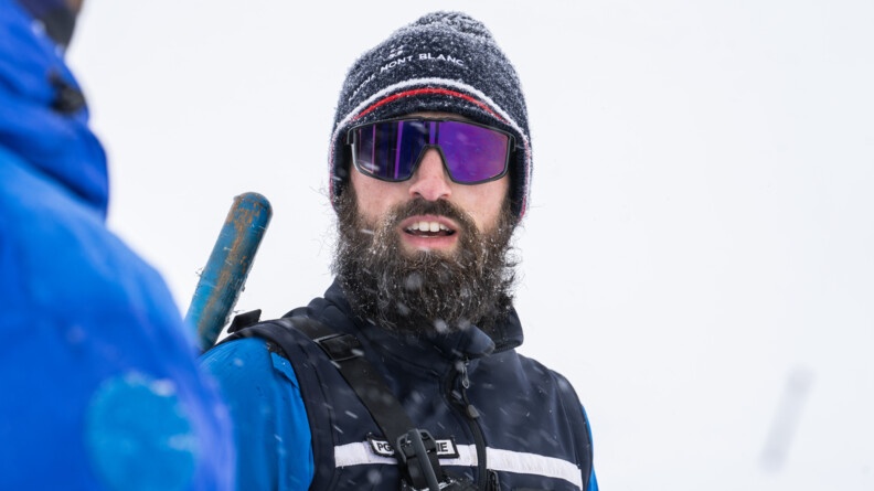 Agrandir l'image Un jeune gendarme se tient debout face à l'objectif. Il porte un gilet de montagne bleu marine, porté sur un vêtement bleu moyen. Il porte une sacoche en bandoulière que l'on devine, ainsi que l'antenne d'une radio. Il porte une barbe brune, des lunettes de ski violettes ainsi q'un bonnet gris avec des liserets rouge et blanc, sur lequel est inscrit Savoie mont-Blanc. On devine également le manche bleu de la pelle qu'il porte dans le dos, dépassant au-dessus de son épaule droite. Il neige légèrement. La barge et le bonnet reretiennent quelques flocons. A l'extrémité gauche de l'image, on devine un homme de dos, portant également un vêtement de neige bleu moyen, tourné vers le gendarme. L'arrière plan est tout blanc, sans aspérité et légèrement flouté.