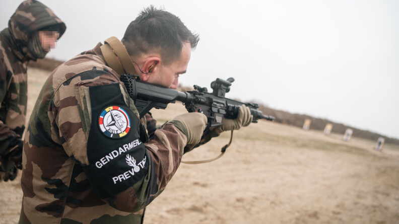 Agrandir l'image De trois quart arrière droit, un prévôt arborant au bras droit le brassard noir de la gendarmerie prévôtale s'entraîne au tir avec une arme d'épaule, sur un terrain sableux. Derrière lui, un militaire le guide