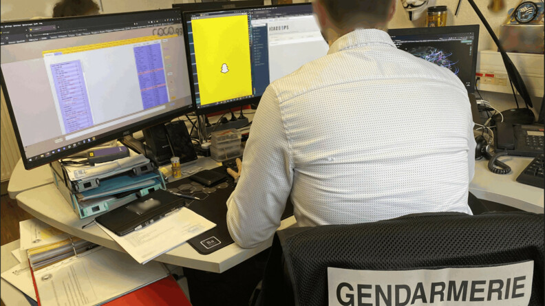 Agrandir l'image Un enquêteur de dos à son bureau devant trois écrans, l'écran du milieu ouvert sur le site Snapchat; sur le siège du gendarme un bandeau écrit en noir sur fond blanc "gendarmerie section de recherches"