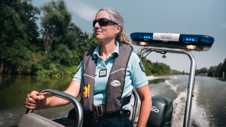 Une femme gendarme se tient debout, sur une embracation légère, avançant sur un fleuve. Equipée d'un gilet de sauvetage et de lunettes de soleil noires, elle se tient à l'armature métllique du siège situé devant elle.