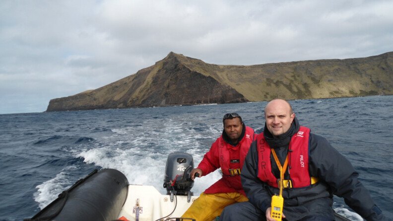 Deux hommes sur un petit bateau devant une ile sans habitation.