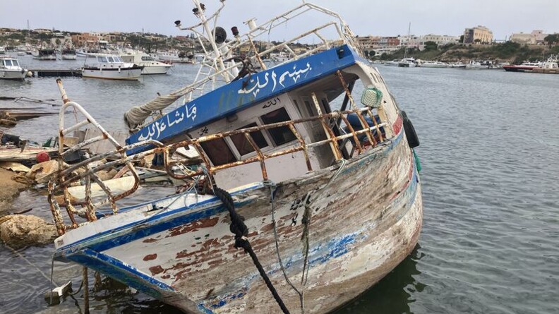 Agrandir l'image Un bateau en très mauvais état, dont la peinture s'écaille, échoué dans les rochers, à l'entrée d'un port. En arrière plan, des bateaux dans le port et des immeubles sur le littoral..