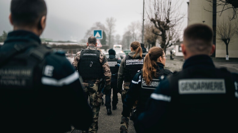 Un groupe de gendarmes marchent dans une rue, bordant des immeubles. Le ciel est gris et peu lumineux.