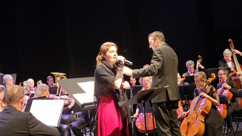 Agrandir l'image La gendarme Stéphanie, alias Lina LoEv, vêtue d'une robe rouge et d'une étole noire, chantant sur scène accompagnée par un orchestre philharmonique.