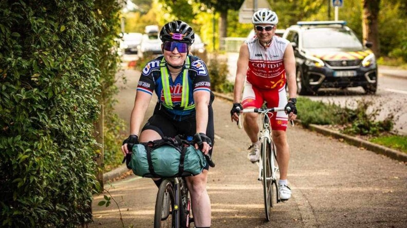Agrandir l'image Une jeune femme cycliste circule sur son vélo. Elle porte un casque et des lunettes. Derrière elle, un homme circule également en vélo. En arrière plan, sur la route, circule une voiture de gendarmerie.