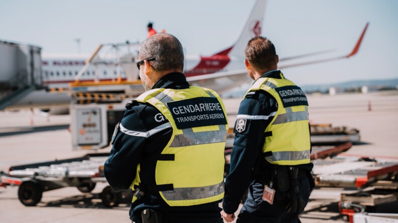 Deux gendarmes vêtus d'un gilet jaune chasuble, porté au-dessus leur uniforme bleu marine, marchent sur le tarmac d'un aéropport. Autour d'eux, au sol, se trouvent toutes sortes d'engins métalliques équipés de roulettes. En arrière plan, on distingue un avion de ligne de couleur blanche et rouge.