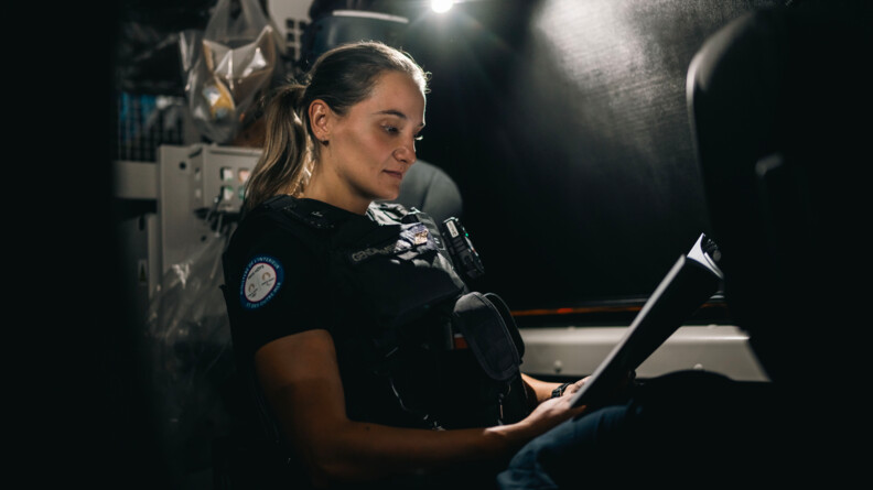 Une jeune femme gendarme, vêtue d'une tenue noire et d'un gilet par-balles, se tient dans un véhicule. Elle observe un document, qu'elle tient dans ses mains.