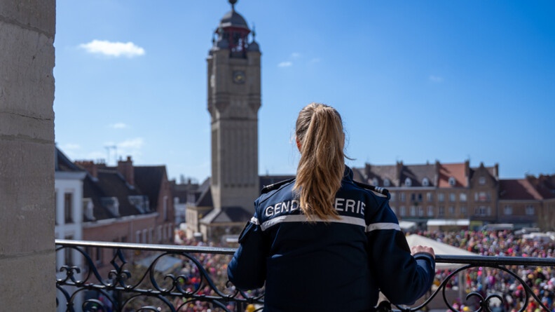 Une femme gendarme, blonde avec une queue de cheval, se tient debout au balcon de la mairie de Bergues, face au Beffroi. Le ciel est bleu et on aper&ccedil;oit les carnavaleurs d&eacute;guis&eacute;s devant la mairie