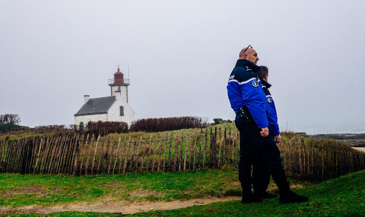 Agrandir l'image Au premier plan à droite de profil, deux gendarmes. Au second plan le littoral breton, recouvert d'herbe et bordé de petites barrières et bois. Au troisième plan, vers la gauche, une maison bretonne blanche au toit d'ardoises devant un phare au sommet rouge. Le tout surplombé d'un ciel brumeux et gris