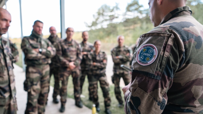 A droite, de dos , un gendarme en treillis arborant sur son bras gauche l'écusson de la F2NRBC de la gendarmerie nationale. Face à lui, flou, 7 militaires en treillis debout l'écoutent attentivement