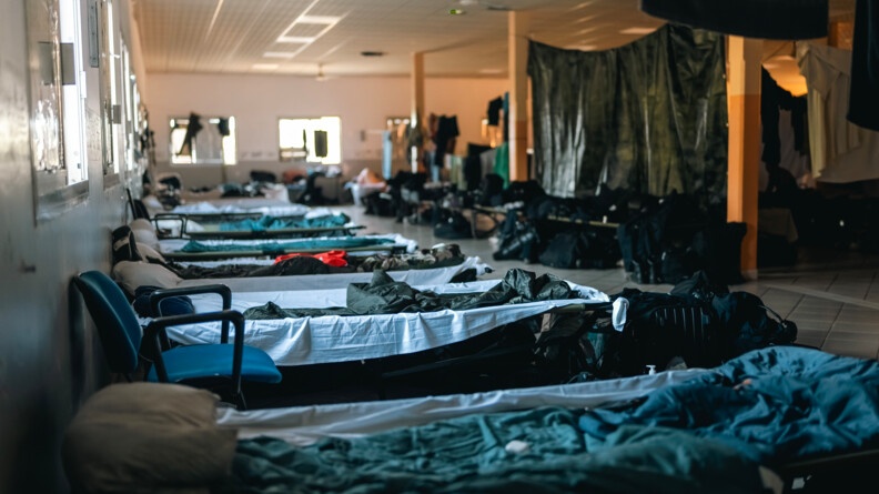 Vue sur une rang&eacute;e de lits de camps install&eacute;s c&ocirc;te &agrave; c&ocirc;te dans une grande salle.