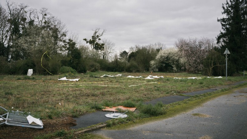 Paysage du lieu de l'exercice. Au sol des zones délimitées par des rubans de gendarmerie. Une dizaine de draps mortuaires au sol pour illustrer le corps des victimes. Tout cela est situé dans de l'herbe, un peu boueuse et humide.