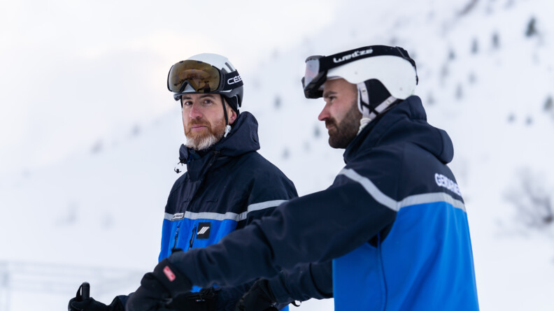 Agrandir l'image Deux hommes, en tenue de gendarmes, posent à l'arrêt sur des skis. Tous deux portent un casque et des lunettes. Ils se tiennent de biais. L'un deux est tourné vers l'objectif, qu'il regarde. Derrière eux, on devine la montagne enneigée, qui est floutée