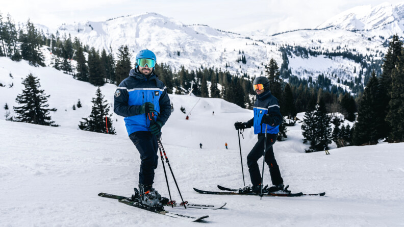 Agrandir l'image Deux gendarmes en ski posent en haut d'une piste enneigée. Equipés d'un casque et d'un masque, tous deux regardent l'objectif en souriant.