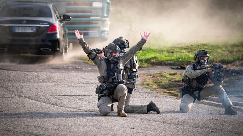 Agrandir l'image Trois hommes du GIGN, armés de fusil, accroupis au sol. Deux sont face à nous. Le troisième leur tourne le dos. En arrière plan, on aperçoit un véhicule noir Mercedes.