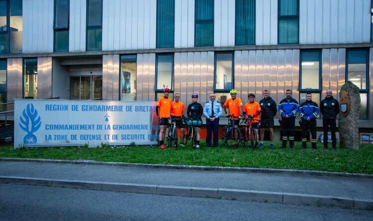Agrandir l'image Une jeune femme cycliste, pose entourée de deux cysclistes avec un t-shit ornage sur sa droite, deux autres cyclistes également avec un t-shit ornage sur sa gauche, ainsi que cinq autres personnes (gendarmes). Derrière eux se trouve un bâtiment, et sur leur droite un grand panneau portant l'inscription Région de gendarmerie de Bretagne