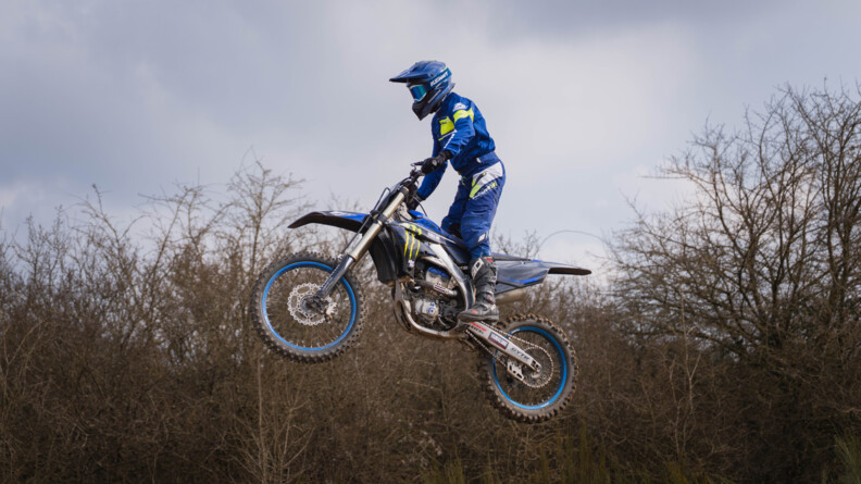 un motard de la gendarmerie sur une moto cross bleue, dans les airs, vu de profil, devant des arbustes