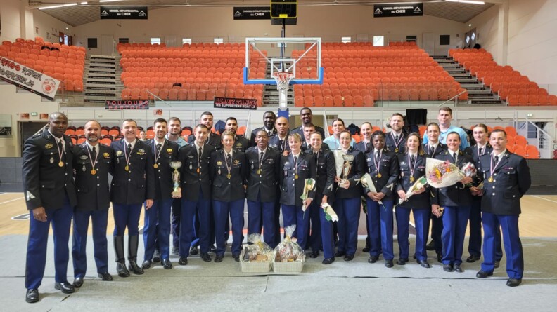 Les équipes de basket de la gendarmerie nationale, masculine et féminine, posent sur un terrain de basket devant le panneau et les tribunes vides.